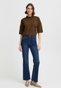 Braun strukturierter Oberteil mit einem Stehkragen, kombiniert mit blauen Denim-Jeans im geraden Schnitt und beigen Ballerinas, präsentiert ein lässiges Outfit.