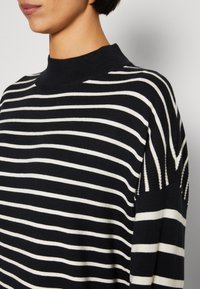 Mustavalkoinen raidoitettu neulepusero, jossa on korkea kaula-aukko ja rento istuvuus. Huomattavat ribbireunat olkapäillä, joissa on vaihtelevaa raitaleveyttä.