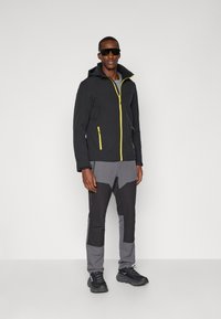 Svart softshell-jakke med gule glidelåser og hette; grå flerfargede bukser med vinklede fargeblokker; svarte joggesko; mørke solbriller.
