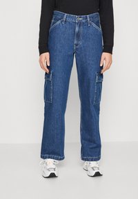 Blå denim cargobyxor med en lös passform, sidofickor och knappstängning; matchade med vita och grå sneakers.