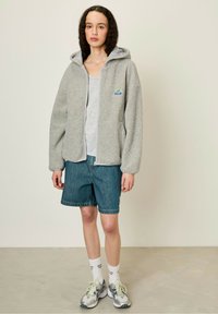 Siva flis zip-up kapuca z mehkimi teksturami, velika kapuca in nalepka na levem prsnem delu, kombinirana z jeans kratkimi hlačami in supergami.