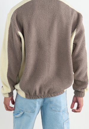 Person trägt zweifarbige Fleecejacke in Braun und Beige mit langen Ärmeln und hellblauen Jeans, von hinten gezeigt.
