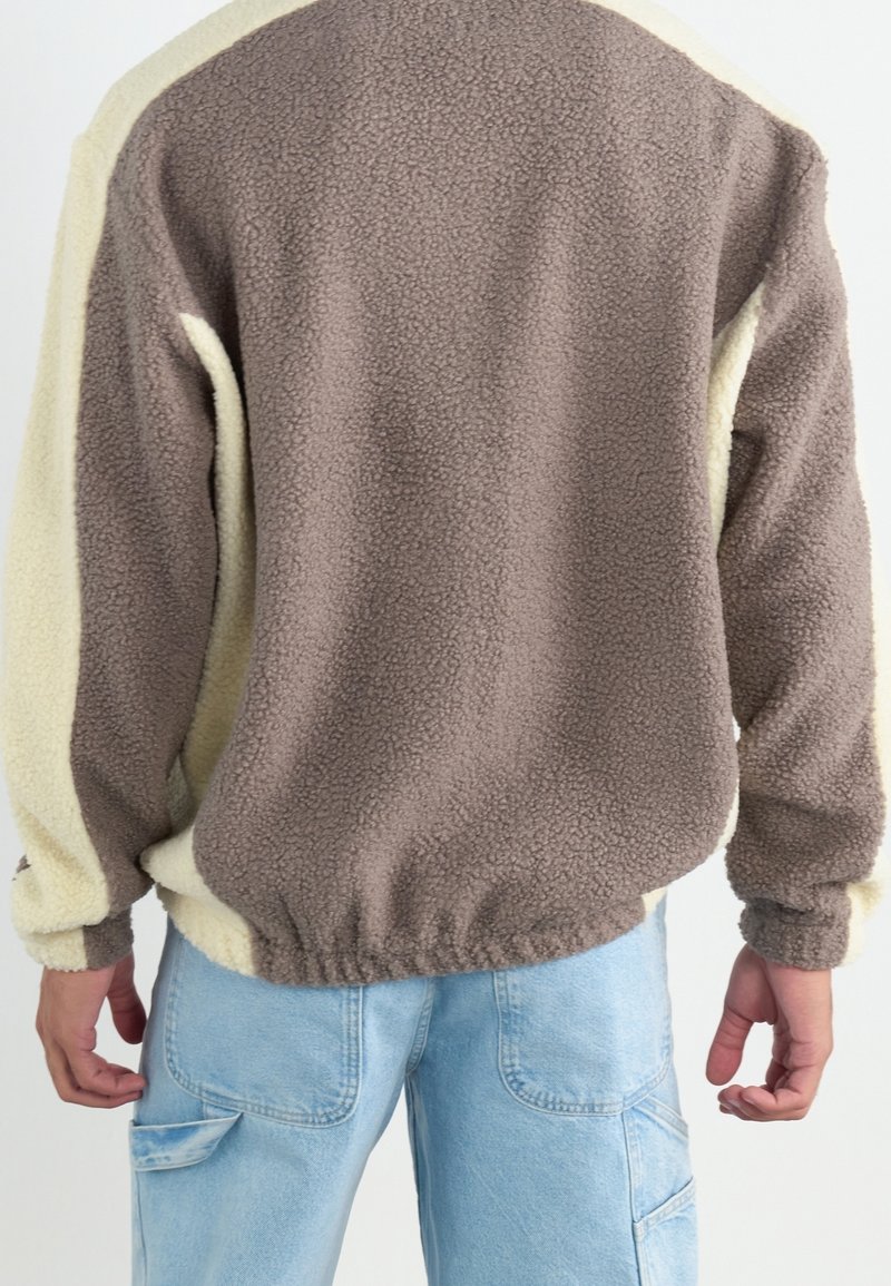 Personne portant une veste polaire bicolore marron et beige à manches longues et un pantalon en jean bleu clair, vue de dos.