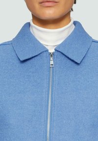 Polarowy koczek w jasnoniebieskim kolorze z zapięciem na zamek z przodu, rozkloszowanym kołnierzem i gładką teksturą. Nałożony na biały sweter z golfem.