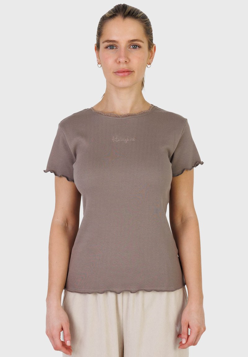 Femme à la peau claire et aux cheveux blonds attachés, portant un haut taupe côtelé à manches courtes avec bords festonnés et un pantalon plissé crème, debout, droite.