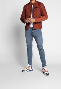 Braune gesteppte Jacke mit hohem Kragen, Reißverschlusstaschen; kombiniert mit blauen Jeans mit Rissen und mehrfarbigen Turnschuhen.