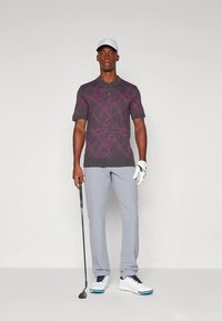 Polo cinzento de manga curta com padrão abstrato roxo, calças cinzentas, sapatos de golfe brancos e um boné branco, segurando um taco de golfe.