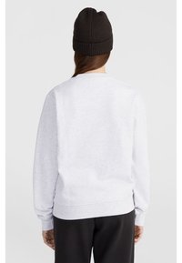 Mujer con un sudadera gris claro, pantalones negros y un gorro de punto marrón oscuro, de espaldas contra un fondo blanco sencillo.