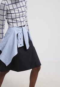 Haut à manches longues rayé gris avec un motif en grille bleu marine, associé à une chemise bleu clair nouée à la taille et une jupe noire jusqu'aux genoux.
