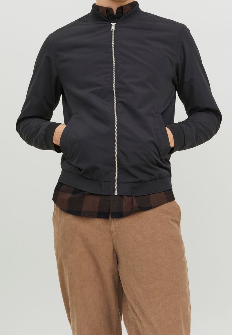 Homme portant une veste bomber noire zippée par-dessus une chemise à carreaux marron et noir avec un pantalon en velours côtelé beige, les mains dans les poches de la veste.