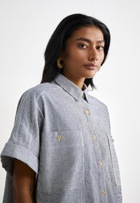 Camicia a righe blu navy e bianca, con due taschini frontali e bottoni dorati. Tessuto morbido con maniche corte e design con colletto.
