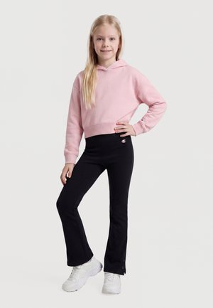Jeune fille debout, souriante, main sur la hanche, portant un sweat à capuche rose, des leggings évasés noirs avec logo et des baskets blanches sur fond neutre.