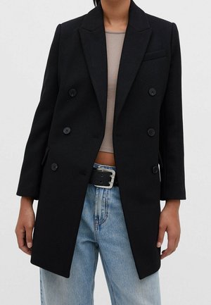 Personne portant un manteau noir croisé sur un haut court beige et un jean bleu clair taille haute avec une ceinture noire.
