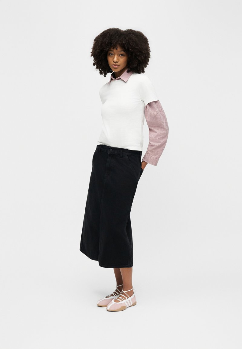 Donna con capelli ricci che indossa una maglietta bianca a maniche corte sopra una camicia rosa a maniche lunghe, culotte a gamba larga nere e ballerine rosa con cinturini.