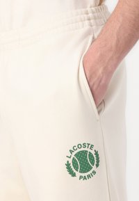 Beige sweatpants med elastisk talje og lommer. Har et grønt Lacoste-mærke med et tennismotiv på venstre ben.