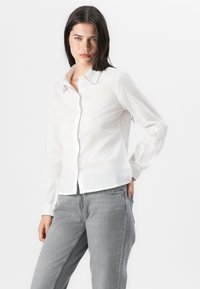 Una mujer se encuentra de pie con confianza, vistiendo una camisa blanca de botón con cuello y jeans grises, frente a un fondo blanco liso.
