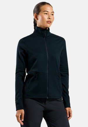 Woman wearing a black zip-up jacket with high collar and zippered side pockets, paired with black pants, standing against a white background.
