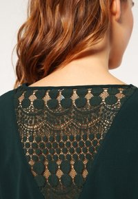 Blouse vert foncé avec un dos en dentelle décorative présentant des détails circulaires et un motif travaillé, alliant un tissu texturé à l'avant lisse.