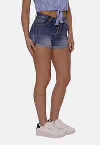 Shorts in denim di un blu medio con effetto sbiadito, caratterizzati da una chiusura a cinque bottoni, orlo arrotolato e due tasche frontali. Abbinati a sneaker bianche.