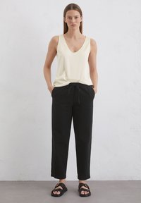 Crème-kleurig mouwloos topje met een V-hals, gecombineerd met zwarte broeken met een trekkoord. Het model draagt zwarte sandalen met een accent.