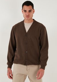 Cardigan marrone con scollatura a V, maniche lunghe e chiusura con bottoni. Realizzato in tessuto morbido con polsini e fondo a costine. Presenta una vestibilità rilassata.