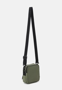 Kompaktowa oliwkowo-zielona kwadratowa torebka na przekór z czarnym regulowanym paskiem i zapięciem na zamek, zawieszona na białym tle.
