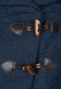 Dettaglio di un cappotto di lana blu navy con accenti in pelle, toggles in legno e lacci intrecciati. Finitura testurizzata con elementi hardware lisci.