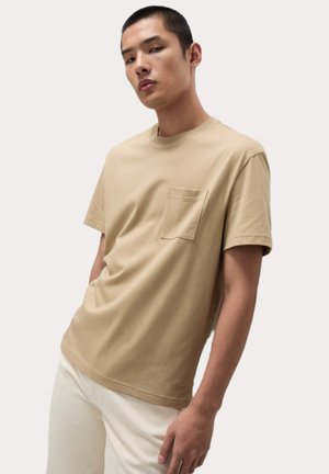 Beige katoenen t-shirt met een ronde hals en korte mouwen, voorzien van een voorzak met een gestikte rand, gedragen met lichtgekleurde broek.