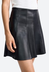 Fustă din piele neagră cu un design evazat, având cusături verticale, textură netedă și lungime până la mijlocul coapsei. Mână odihnindu-se pe talie.
