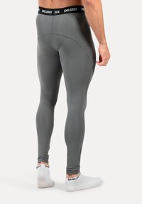 Man in grijze compressieleggings en witte enkelsokken met zwart "Smilodox"-merk, staand met zijn rug en rechterarm zichtbaar.