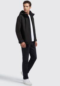 Giacca impermeabile nera con cappuccio, zip frontale e polsini regolabili, abbinata a jeans scuri e sneakers bianche. Design semplice e lineare.