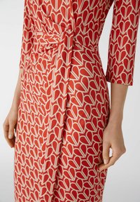 Robe rouge avec un motif géométrique crème présentant des formes allongées ; le tissu est lisse et possède un détail à la taille nouée. Manches à trois-quarts.