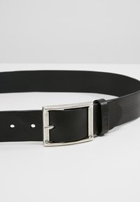 Cinturón de cuero negro con una hebilla rectangular de plata y múltiples agujeros de ajuste. Detalle del logo estampado en la correa.