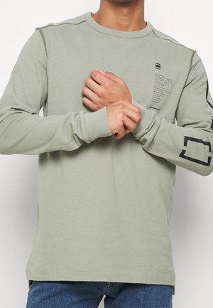 Homme portant une chemise à manches longues vert clair avec texte noir et motif géométrique, ajustant les manches, montré du cou jusqu'aux hanches.