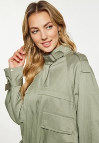 Jachetă verde khaki cu guler înalt, accente cu nasturi și patru buzunare frontale. Textură netedă cu un design structurat și mansete ajustabile.