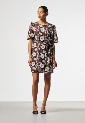 Frau trägt ein knielanges braunes Kleid mit weißer Blumenstickerei und kurzen Ärmeln, steht mit den Händen an den Seiten und schaut nach links.