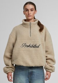 Pullover in pile beige con colletto alto, zip frontale e la parola "Proibito" in corsivo nero al centro. Tessuto morbido, vestibilità comoda.