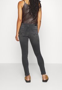 Jeans skinny negros con bolsillos traseros que presentan costuras decorativas, combinados con una blusa de cuero marrón y sandalias planas.