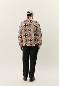 Moški v bež flis jakni z rdečimi in črnimi geometričnimi vzorci, črnih kavbojkah, rjavih čevljih in bež kapo, stoji s hrbtom proti gledalcu.