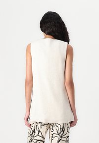 Sleeveless beige linen top with a straight cut, featuring a high neckline and side slits, paired with patterned black and white trousers.