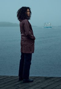 Waterdichte, kastanjebruine mid-length jas met een capuchon, gedragen over donkere jeans en zwarte laarzen, staat tegen een mistige waterkant achtergrond.