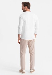 Mies, joka pitää yllä valkoista pitkähihaista paitaa, beigejä housuja ja beigejä lenkkareita, seisoo selkä kameraan valkoista taustaa vasten.