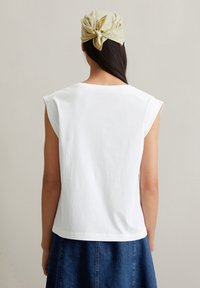 Witte katoen mouwloze top met een iets gebogen zoom. De achterkant toont een beige patroon hoofddoek knotted in de nek.