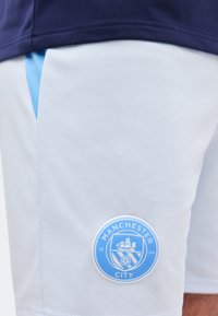Κοντινό πλάνο λευκών αθλητικών σορτς με ανοιχτό μπλε λογότυπο της ποδοσφαιρικής ομάδας Manchester City κοντά στο στρίφωμα και σκούρο μπλε ζώνη μέσης.