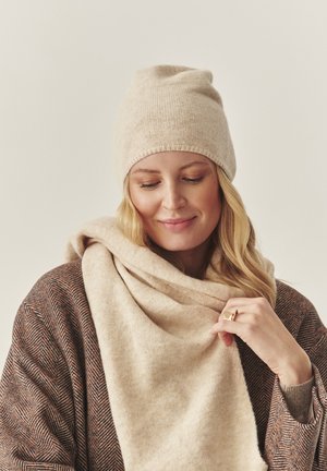 Bežová pletená čepice má těsný střih. Modelka má na sobě světle béžový šál a hnědý cardigan s vzorem, což dodává texturu a teplo.