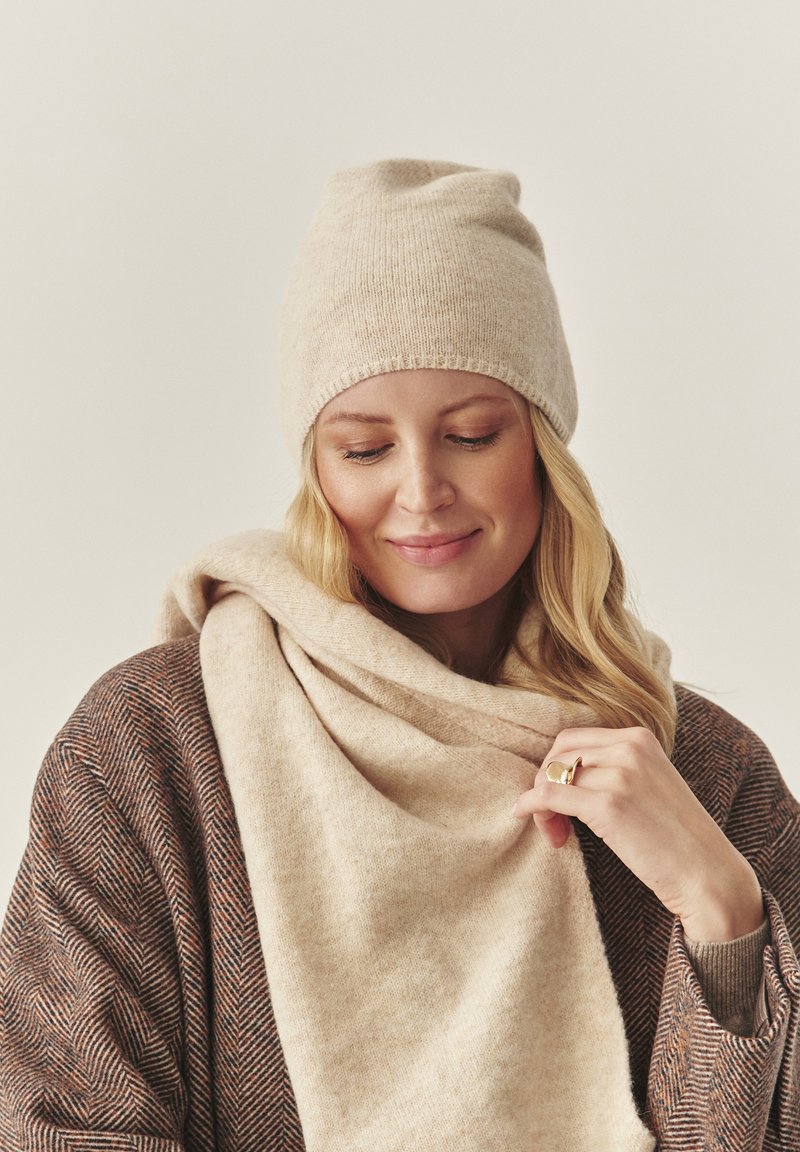 Béžová pletená čiapka má priliehavý strih. Modelka nosí svetlobéžový šál a hnedý vzorovaný kardigán, ktorý pridáva textúru a teplo.