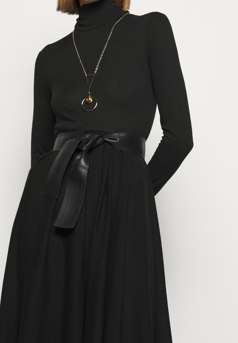 Robe col roulé noire à manches longues, cintrée à la taille par une large ceinture en cuir noir plate avec un détail noué, accompagnée d'un collier pendentif doré.