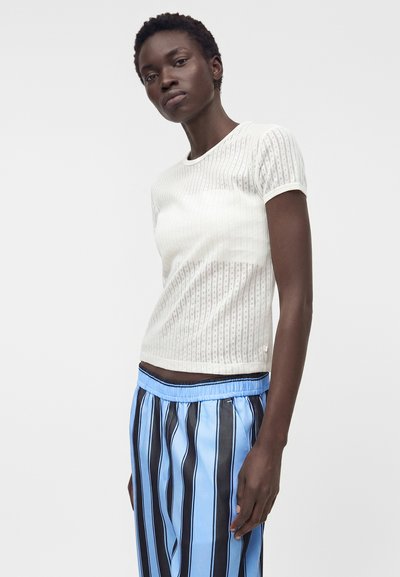 Γυναίκα που φοράει λευκό κοντομάνικο υφασμένο πουλόβερ και φαρδιά μπλε παντελόνια με μαύρες κάθετες ρίγες, στέκεται μπροστά σε μια απλή φόντο.