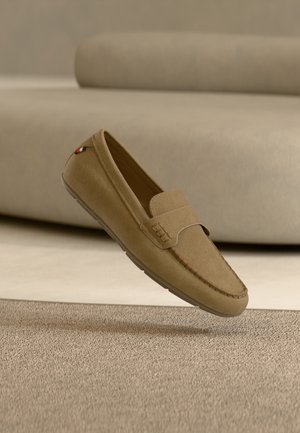 Vaaleanruskea mokkainen loafer, jossa on ommellut yksityiskohdat ja pieni kangasmerkki lähellä kantapäätä, leijuu beigenvärisen maton yllä.