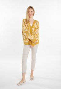 Cardigan giallo senape con motivi floreali bianchi, chiusura a bottoni, vestibilità ampia. Abbinato a pantaloni cropped beige chiaro e scarpe flat.